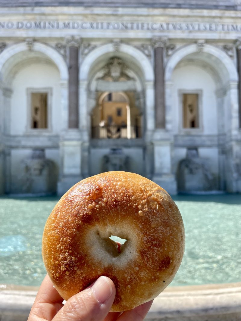 A sesame bagel held up in front of a Roman fountain